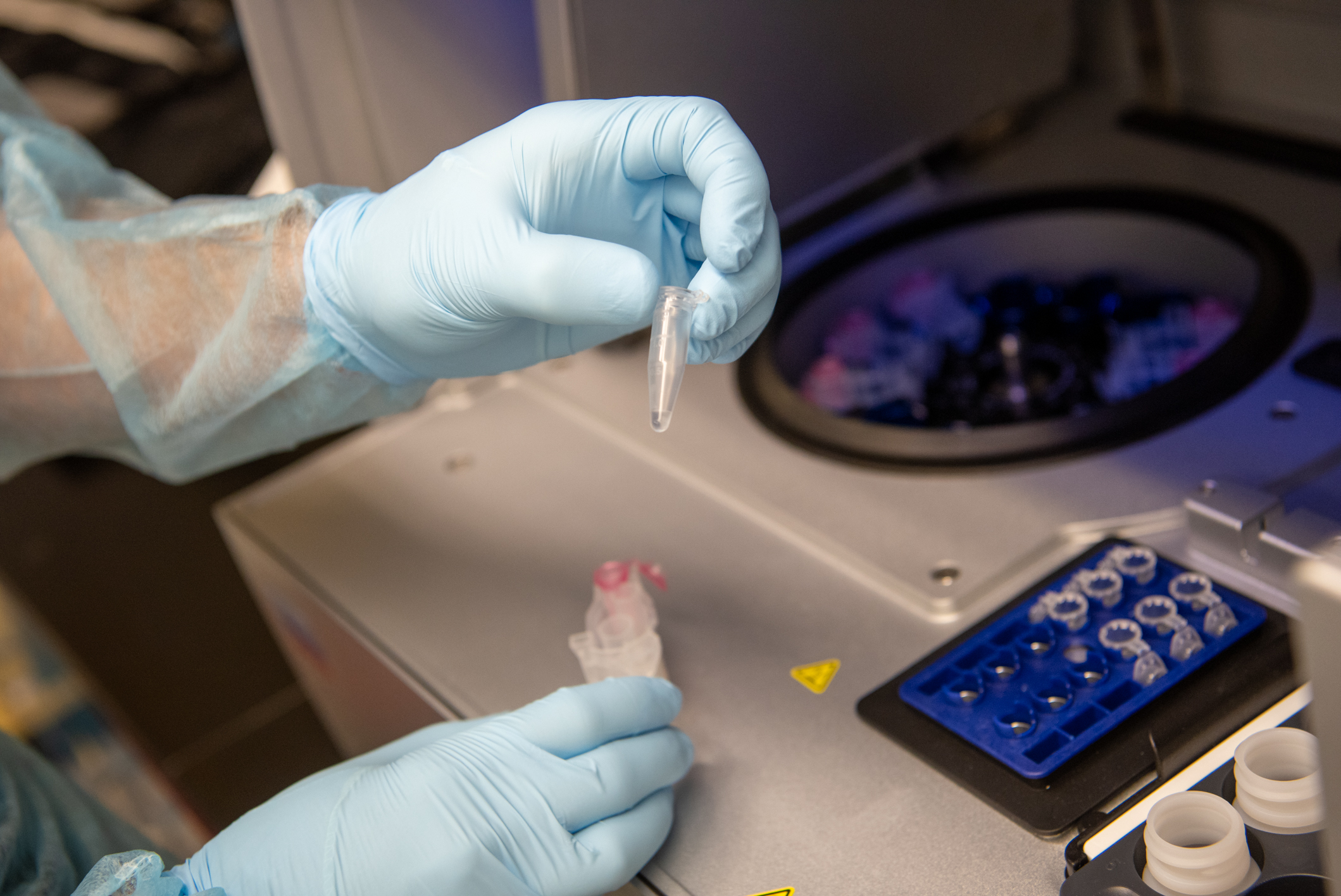 A hand holds a microcentrifuge tube next to the microcentrifuge equipment, with a tray for the tubes beside it.