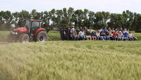 people on a long trailer in a field listening to a speaker at field days