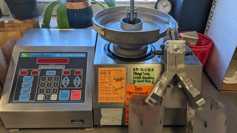 A stainless steel Seedburo counter with a vibrating metal bowl feeds kernels across electronics that count them before dumping into catch basins.