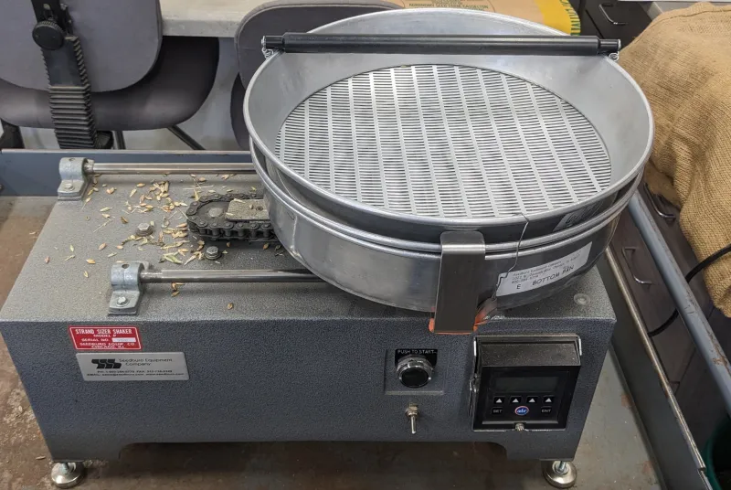 A metal-cased tabletop machine with slotted screens and a round catch basin, where a shaker rotates pans for sorting same-sized seeds.