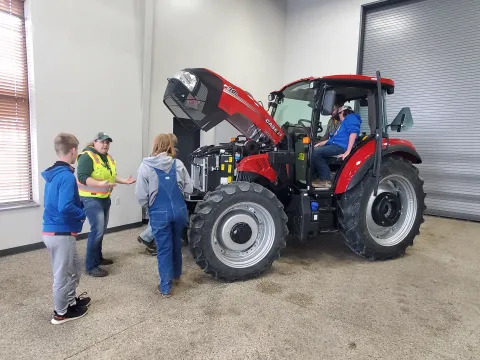 Group observing a red tractor with its hood open; two people sit in the cab